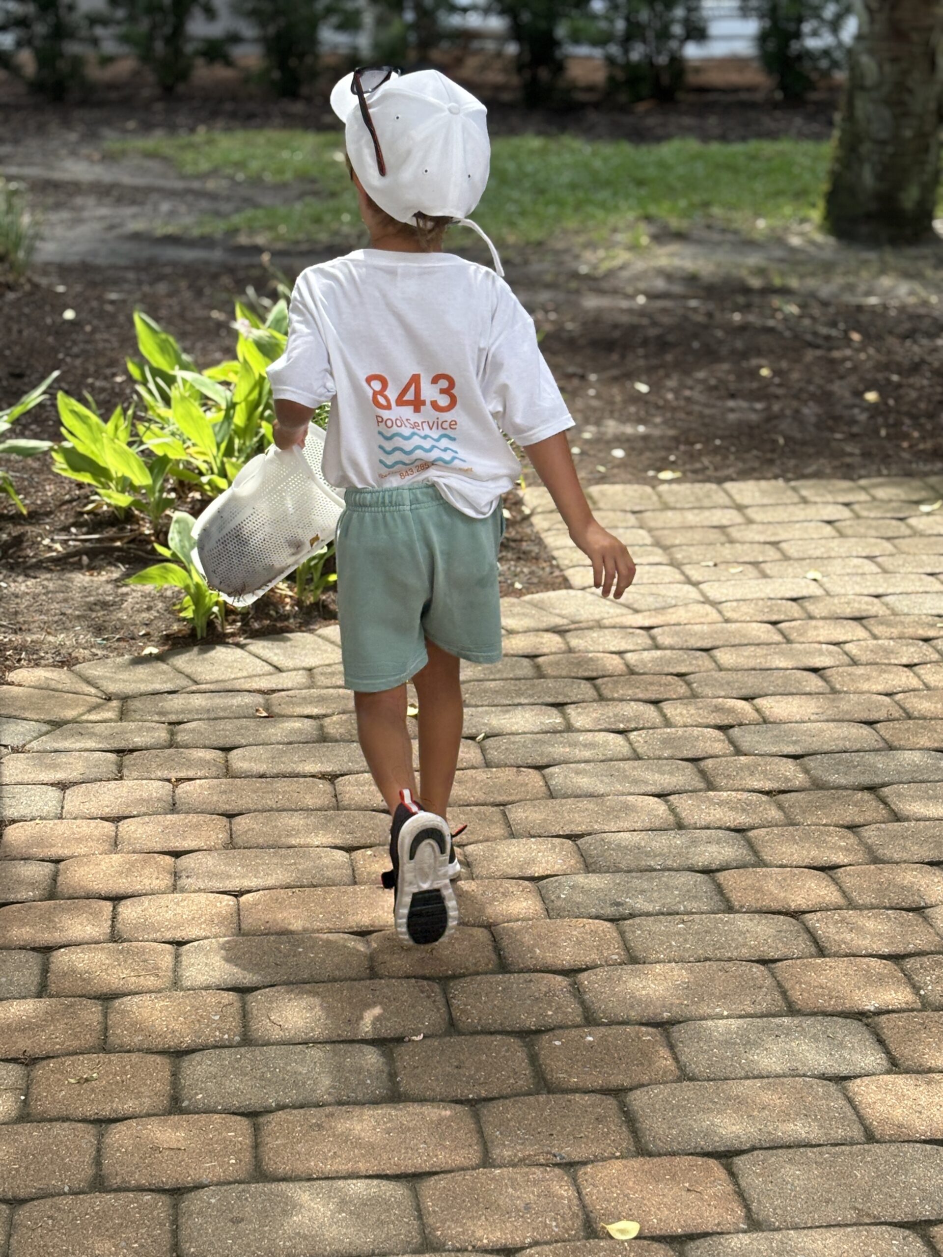 Child in a white sun hat and T-shirt with '843 Poolservice' on the back walks along a brick pathway, carrying a white basket outdoors.